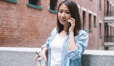 Kot ceketli Asyalı genç kadın telefonda konuşuyor ve korkuluklara yaslanıp kameraya bakarken limonata içiyor.