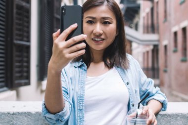 Modaya uygun kot ceketli güzel bir kadın selfie çekip limonata içerken taş parmaklıklara yaslanıp gülümsüyor.