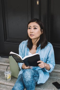 Kot pantolon giymiş, siyah kapılı merdivenlerde oturmuş kitap okurken şaşırmış bir şekilde yukarı bakan etnik bir kız öğrenci.