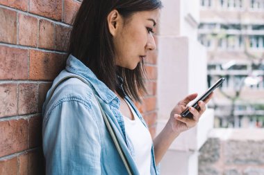 Tuğla duvara yaslanmış bir fincan kahve ve sosyal medyada sohbet eden konsantre ve odaklanmış bir etnik kadının yan görüntüsü