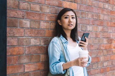 Tuğla duvara yaslanmış, mesaj atarken ve kahve içerken kameraya bakan gündelik kıyafetlerle güzel, esmer bir kadın.
