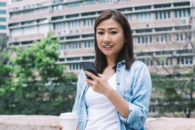 Tuğla çitin yanında dikilmiş elinde kahve tutarken kameraya bakan ve akıllı telefon kullanan mutlu, güler yüzlü etnik kadın.