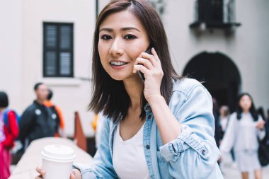 Gündüz vakti şehir sokağında siperlere yaslanırken elinde kahve fincanı olan modern Asyalı kadın akıllı telefonla konuşuyor.