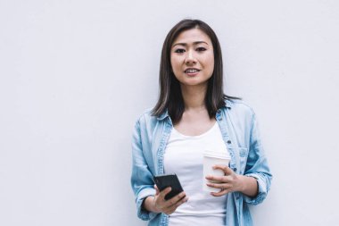 Genç Asyalı kadın mavi gömlek ve kot pantolon giyiyor. Akıllı telefonuyla duvara yaslanıyor ve kameraya bakarak kahve alıyor.