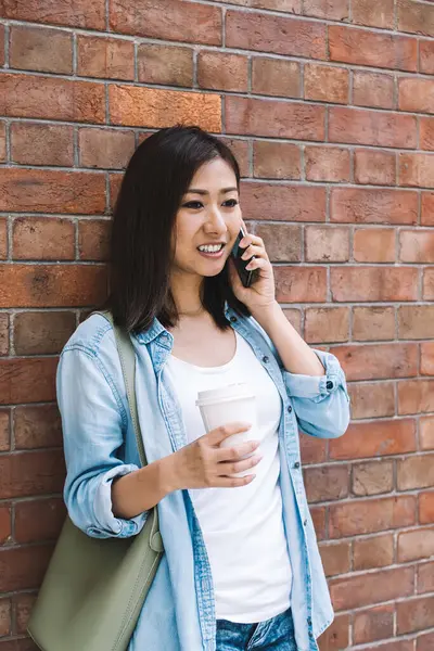 Kot pantolon giymiş kendine güvenen Asyalı kadın telefon görüşmeleri sırasında tuğla duvarın yanında kahve ve akıllı telefonla duruyor.