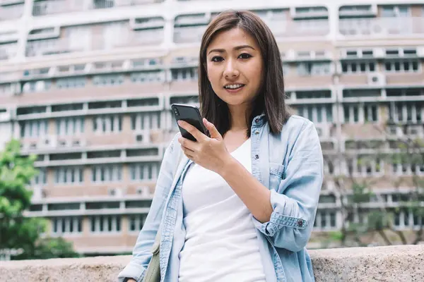 Tuğla çitin yanında durup kahve içerken kameraya bakan ve telefon kullanan gündelik giysiler içinde gülümseyen etnik bir kadın.