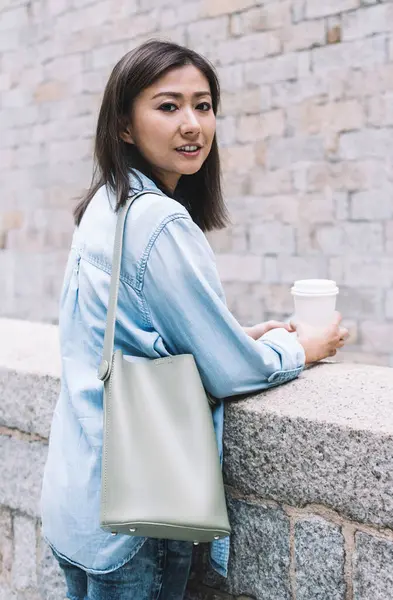 Tasasız, genç Asyalı kadın, güpegündüz sokakta tuğladan korkuluğun yanında durup gülerken kahve fincanı alıp gülümsüyor.