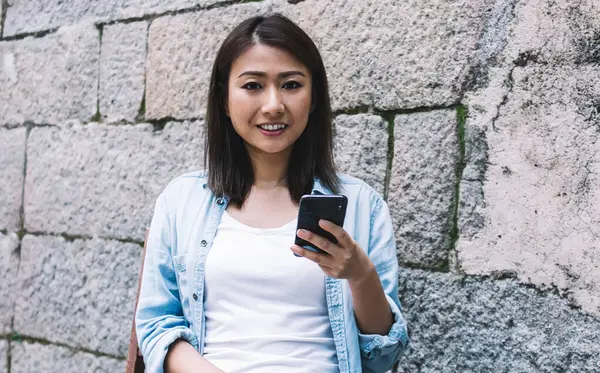 Havalı, neşeli, limonata bardağında limonatayla gezen, akıllı telefonuyla sokakta, tuğla duvarın yanında otururken mesaj atan genç Asyalı bir kadın.