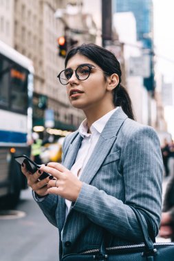 Ciddi kadın çalışanların yan görüntüsü yuvarlak gözlüklü, trend çantalı ve akıllı telefonlu ve başka tarafa bakan.