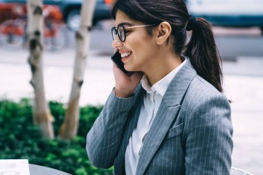 Resmi giyinmiş, gözlüklü, at kuyruklu, tablet ve kafeteryanın dışındaki diyagramlarla cep telefonundan konuşan gülümseyen yetişkin bir kadının yan görüntüsü.