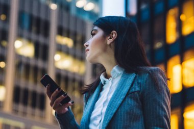 Resmi kıyafet giymiş güzel bir iş kadını, gece şehir merkezindeki modern ofis binalarının önünde dikilirken akıllı telefondan mesajlaşıyor.