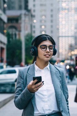 Resmi giyinmiş, yuvarlak gözlüklü, akıllı telefon kullanan ve şehir merkezinde bulanık arka planda yürürken gözleri kapalı müzik dinleyen bir iş kadını.