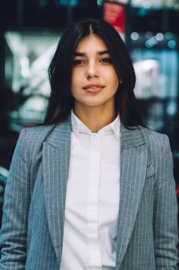 Hoş ve çekici etnik bayan girişimci resmi takım elbiseli uzun siyah saçlı bulanık arka planda kameraya bakıyor.