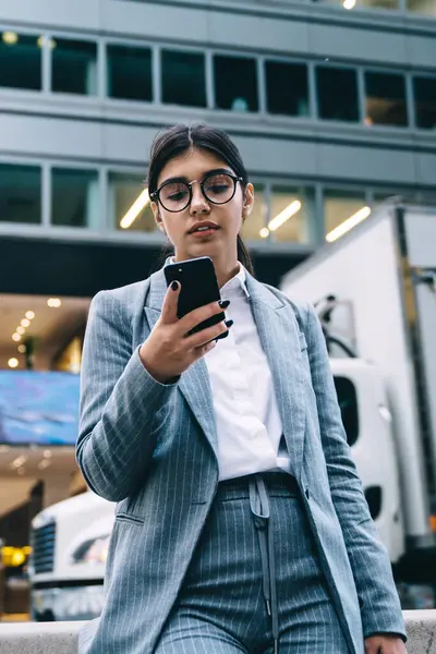 Resmi takım elbiseli ve yuvarlak gözlüklü zeki bayan çalışanı. Akıllı telefonuyla sokakta duruyor ve sosyal ağda mesaj yazıyor.