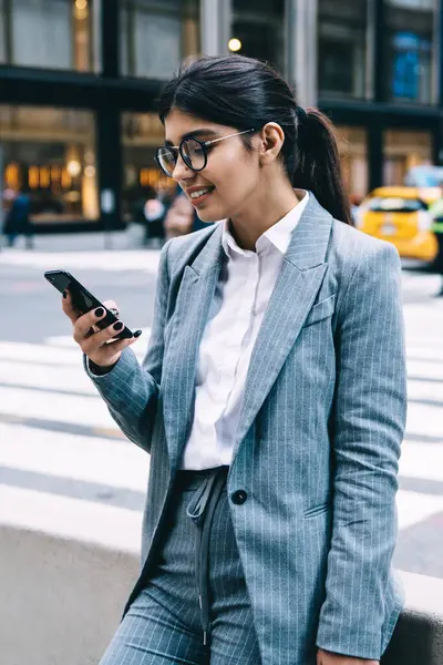 Yuvarlak gözlüklü ve resmi takım elbiseli neşeli genç bayan girişimci akıllı telefonuyla yolun kenarında duruyor ve korkuluklara yaslanırken gülümsüyor.