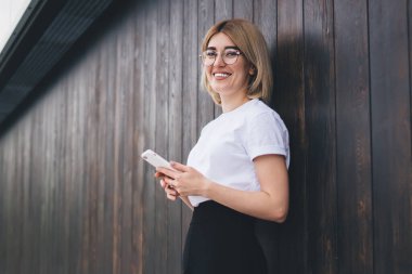Genç, gözlüklü, akıllı telefon kullanan, bisikletin yanında duran, binaların ahşap duvarına yaslanan, neşeyle gülümseyen ve kameraya bakan genç bir kadın.