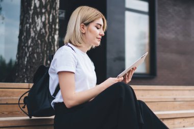 Sokak binasının merdivenlerinde bisikletle otururken siyah sırt çantalı, sörf tabletli yetişkin bir kadının yan görüntüsü.
