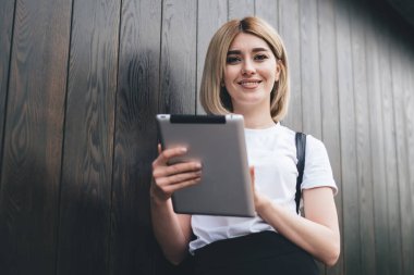 Gün ışığında kameraya gülümseyen, ayakta duran ve tahta duvara yaslanan neşeli, genç bir kadının tablet sörfü sahnesi.