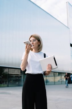 Gözlüklü, sıradan kıyafetler giyen, akıllı telefondan konuşan ve ofis binasının yanındaki caddede dikilirken gözlerini kaçıran düşünceli bir kadın.