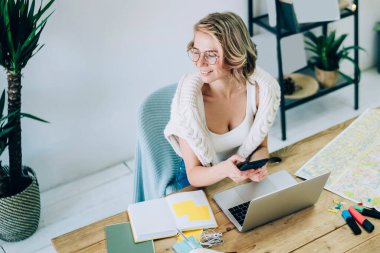 Elinde akıllı telefon cihazıyla mutlu kadın serbest çalışanın uzaktan iş için rahat bir yerde oturduğunu, seyahatleri planlarken yüzünü çevirdiğini ve gülümsediğini görüyoruz.