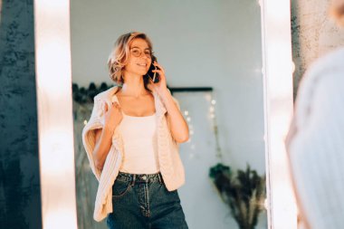 Optik gözlüklü mutlu bir kadın soyunma odasındaki lamba aynasının yanında poz veriyor ve moda tasarımcısıyla cep telefonu konuşması yapıyor. Moda kıyafetlerini tartışıyor, telefonla gülümseyen bir kadın arıyor.