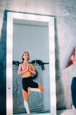 Sağlıklı yaşam tarzı egzersiz konsepti - Kafkas kadın denge yoga yapıyor asana Vrikshasana lamba aynası yanında, eşofmanlı aktif kız ağaçta duruyor ruh ve kapalı alanlarda tam akıl için poz veriyor.