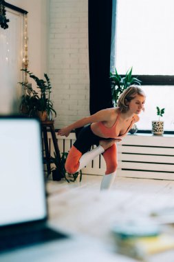 Spor kıyafetleriyle yoga yapan sakin yetişkin bir kadın modern odada güvercin pozu vererek bitki vazolarının arka planında yatağın karşısında duruyor.