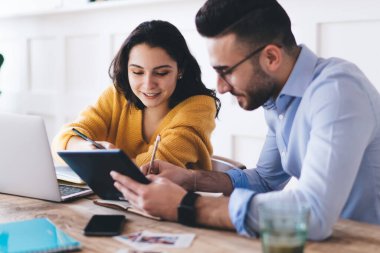 Gülümseyen bir çift, günlük giysiler giyip dizüstü bilgisayara ve tablete bakarken ders kitaplarına not alıyorlar ve evde masada oturuyorlar.