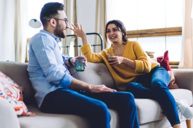 Mutlu genç çift rahat bir koltukta oturup güzel güzel sohbet ederken kadınlar el kol hareketleri yapıyor ve erkekler güneşli odada gülümsüyor.