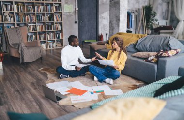 Odaklanmış bir çift yerde oturmuş, rahat bir oturma odasında evrak işlerini tartışıyorlar. Belgelerle çevrelenmiş olarak, takım çalışmalarını ve sorumluluklarını gösteren mali veya işle ilgili planlama konusunda işbirliği yaparlar.