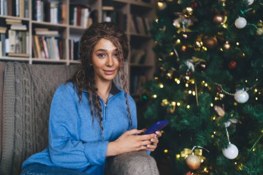 Genç bir kadın süslü bir Noel ağacının yanında akıllı telefon kullanıyor, muhtemelen online para transferi yapıyor. Sıcak tatil ortamı ve şenlikli ışıklar sıcak, neşeli atmosferi güçlendirir.