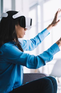 Mavi tişörtlü, uzun siyah saçlı bir kadının VR gözlük takıp sanal duvara dokunuşunun yan görüntüsü.
