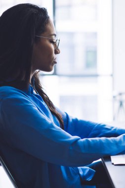 Masaüstünde büyük ekranlı rapor yazarken basılı diyagramlar ve dizüstü bilgisayarla çalışan konsantre genç siyahi kadının yan görünümü