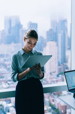 Kendine güvenen güzel bir kadın bilgisayarın yanında dururken dokunmatik ped kullanıyor. Geniş, çağdaş bir ofiste çalışıyor. Şehir manzarası da büyük bir pencerenin arkasında.