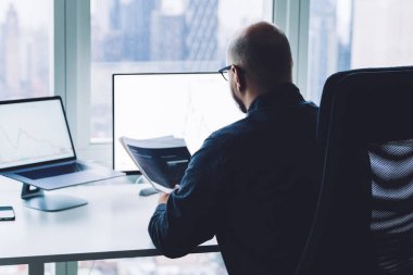 Bilgisayarda çalışan gözlüklü kel adamın arka planda elinde bir yığın kağıt varken ofiste oturduğunu görüyorum.