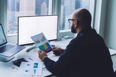 Modern ofiste masa başında oturmuş kağıt grafikleri incelerken bilgisayar ekranına bakan gözlüklü adamın yan görüntüsü