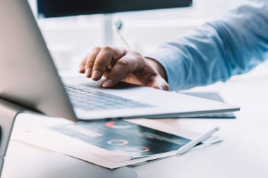 Mavi gömlekli adam ofisteki beyaz bilgisayar masasında oturuyor ve gündüz bilgisayarında çalışırken dokunmatik ped kullanıyor.