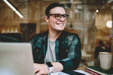 Neşeli hipster erkekte erkek öğrenci uzak yazarak geribildirim online sohbet arıyorsunuz gözlük gülümseyen serbest dizüstü bilgisayar, café iç yazarak yayın makale zevk gözlükler