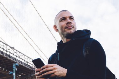 Aşağıdan, kısa saçlı, kıllı, siyah spor giysili, sırt çantalı, akıllı telefonlu genç bir adam. Şehir arka planında, devasa bir köprünün yanında yazılar yazıyor ve başka yöne bakıyor.