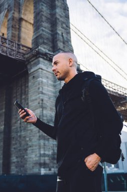 Siyah spor giyim giymiş atletik genç bir adamın akıllı telefonuna tıklaması ve şehir köprüsünün arka planında gün ışığında başka bir yere bakması.