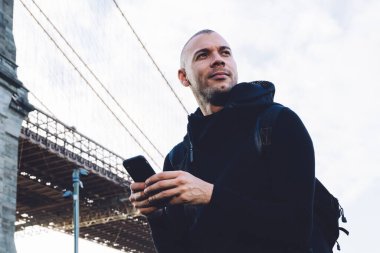 Aşağıdan, kısa saçlı, kıllı, siyah spor giysili, sırt çantalı, akıllı telefonlu genç bir adam. Şehir arka planında, devasa bir köprünün yanında yazılar yazıyor ve başka yöne bakıyor.