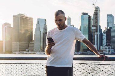 Kısa saçlı, spor elbiseli, gökdelenlerin arka planında duran ve akıllı telefondan ciddi bir yüzle mesaj atan genç bir adam.