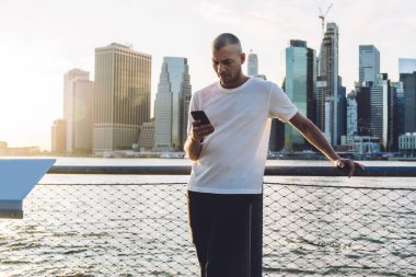 Kısa saçlı, spor elbiseli, gökdelenlerin arka planında duran ve akıllı telefondan ciddi bir yüzle mesaj atan genç bir adam.
