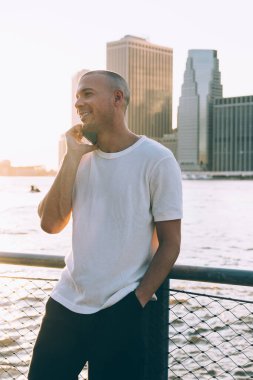 Günlük kıyafetli genç adam suyun yanında duruyor tırabzana yaslanıyor ve telefonda konuşuyor. Yüzü gökdelenlerin arka planında gülümsüyor.