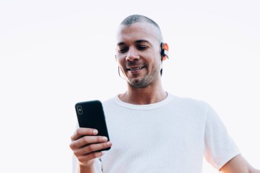 Beyaz gömlekli, kulaklıklı, akıllı telefon kullanan ve şehrin arka planında metalik çit ve nehrin yanında duran genç etnik gülümseyen kısa saçlı adam.