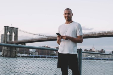 Kulaklıklı ve akıllı telefonlu genç etnik adam modern şehre karşı zincirli metal çitlerin yanında duruyor.