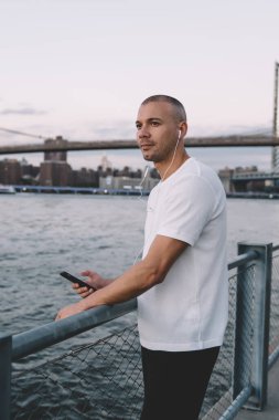 Kısa saçlı, spor elbiseli, akıllı telefon ve kulaklıklı, nehir kenarında zincirli metal çitlere dayanan yakışıklı adamın yan görüntüsü.