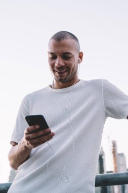 Aşağıdan gülümseyen, beyaz tişörtlü, kulaklıkla müzik dinleyen ve metal çitlere yaslanarak akıllı telefon kullanan genç bir adam.