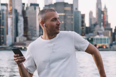 Kısa saçlı, kısa saçlı, kulaklıklı ve akıllı telefonlu yakışıklı genç adam nehir kenarında çitlere yaslanırken gözlerini kaçırıyor.