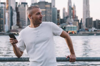 Kısa saçlı, kısa saçlı, kulaklıklı ve akıllı telefonlu yakışıklı genç adam nehir kenarında çitlere yaslanırken gözlerini kaçırıyor.